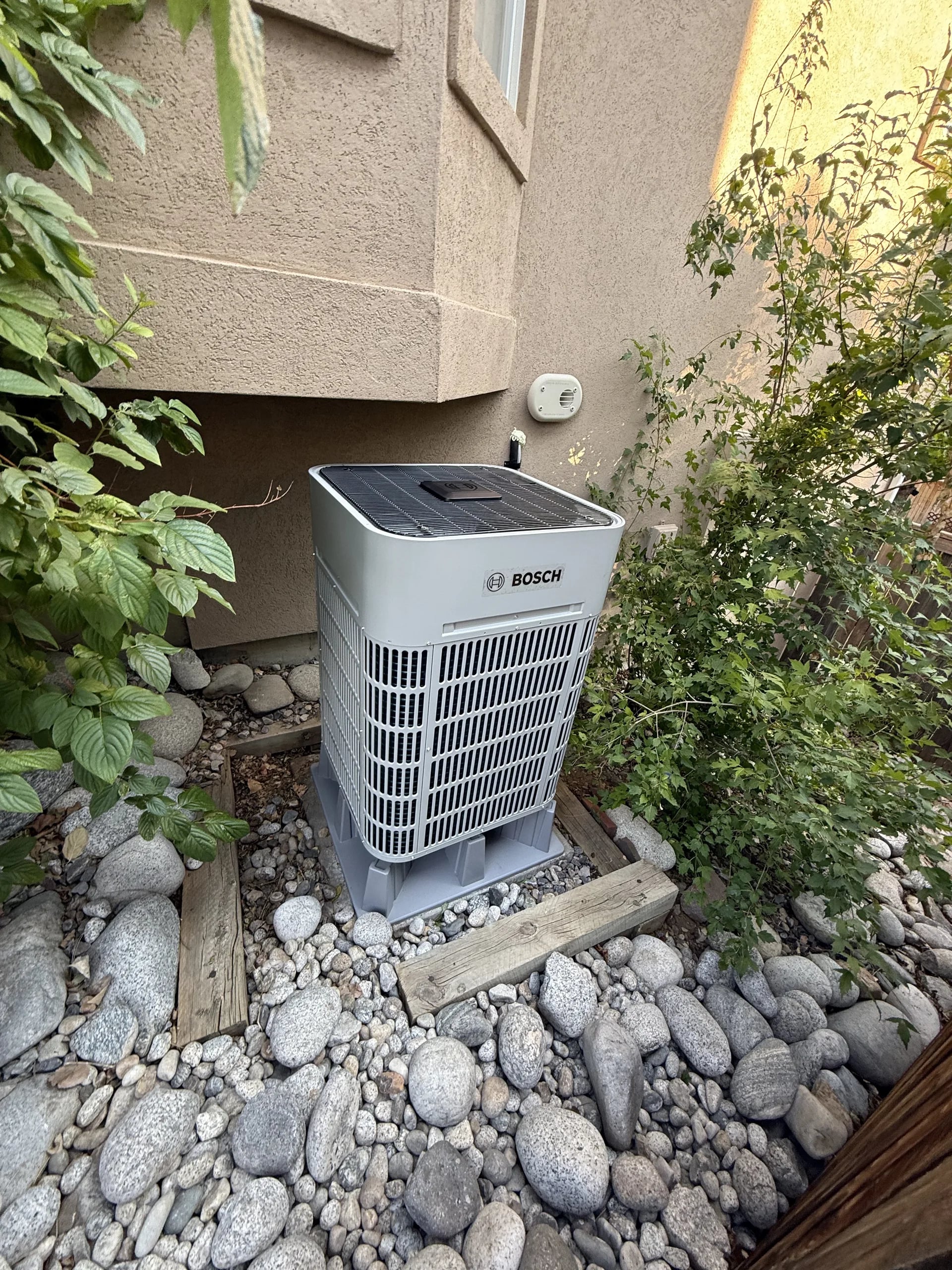 Bosch outdoor unit on risers in a bed of river rock beside a stucco wall