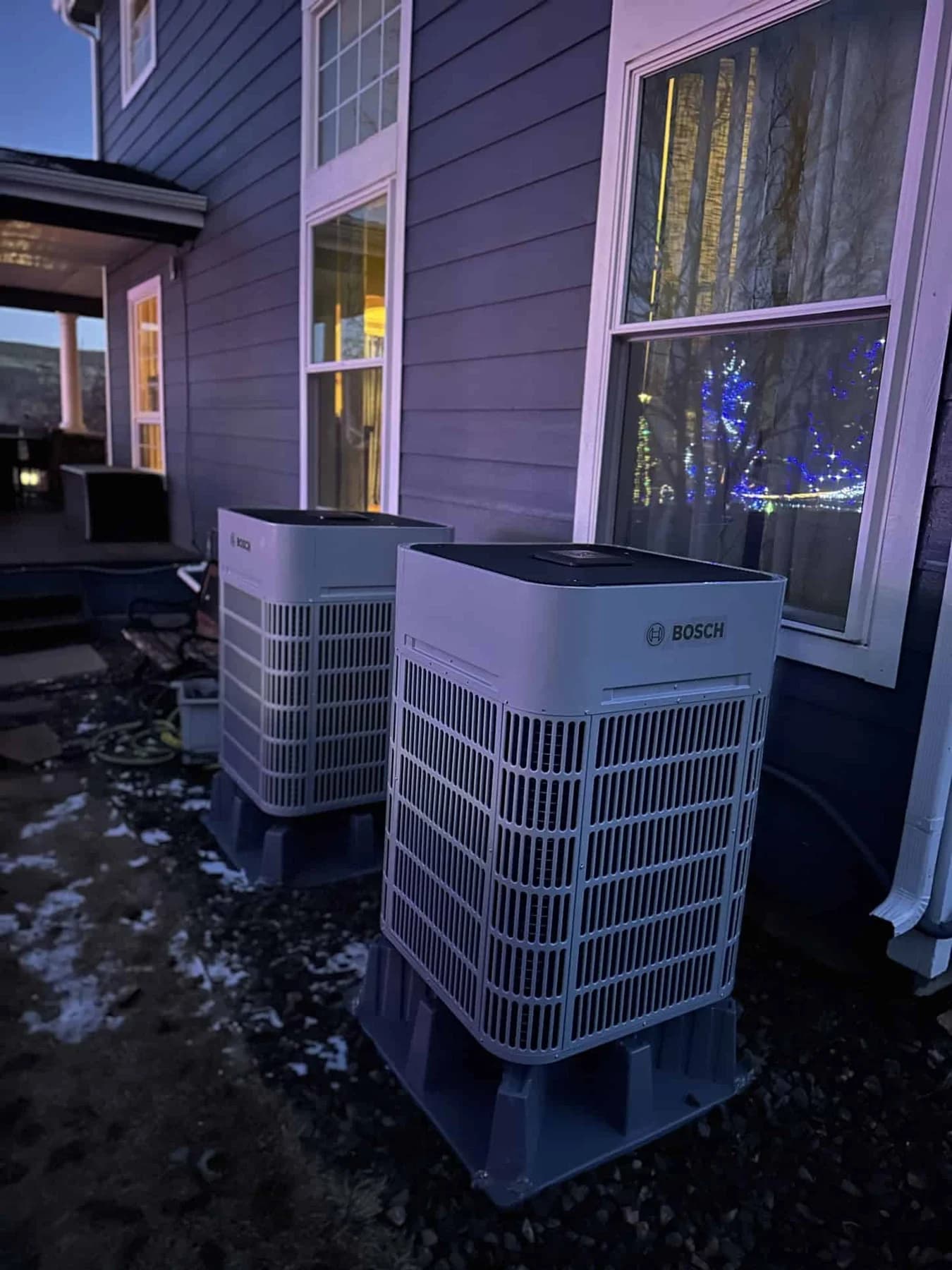 Two Bosch outdoor heat pumps beside a house in snow at dusk